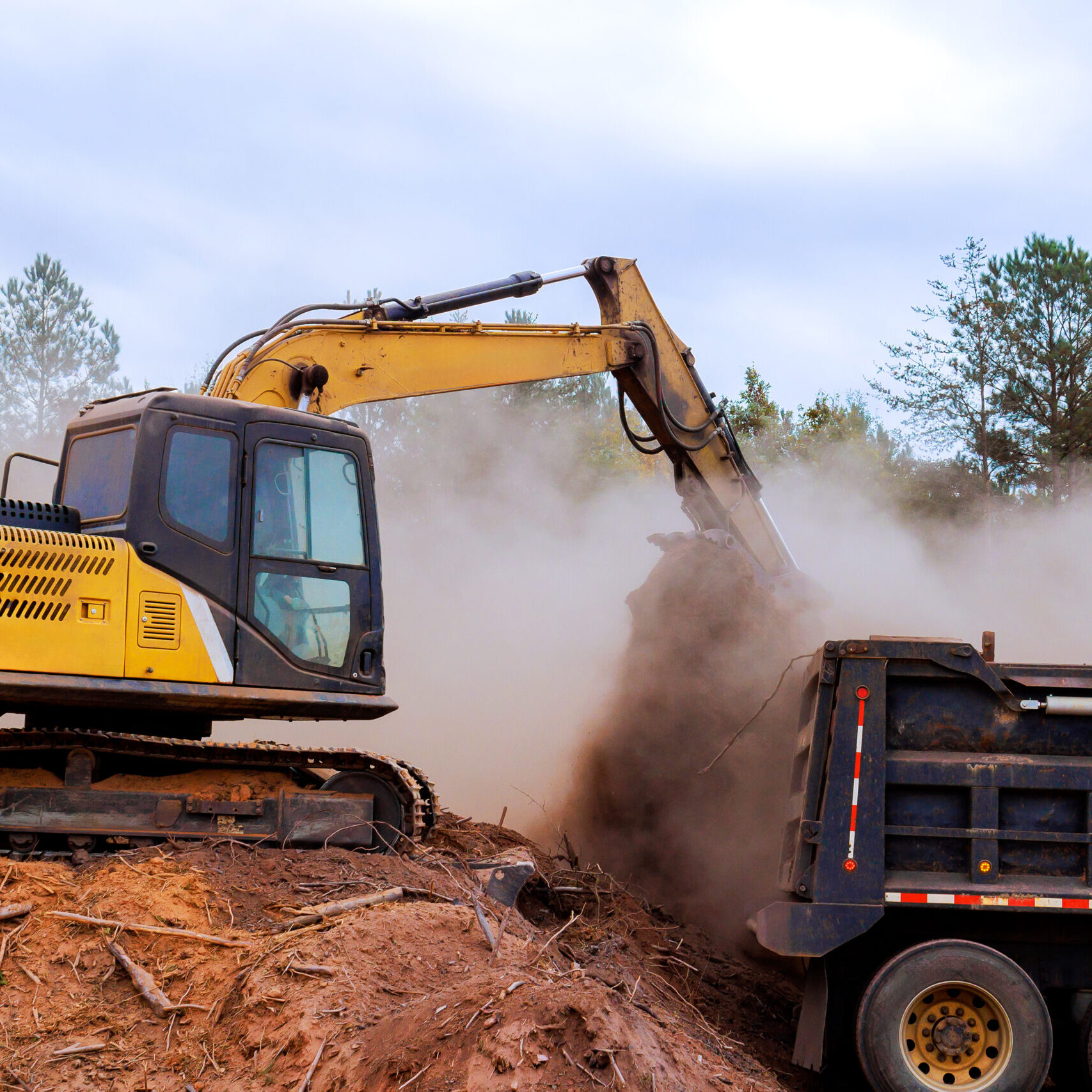 Excavator moves dirt, debris into dump truck amid dust cloud on construction site surrounded by trees.